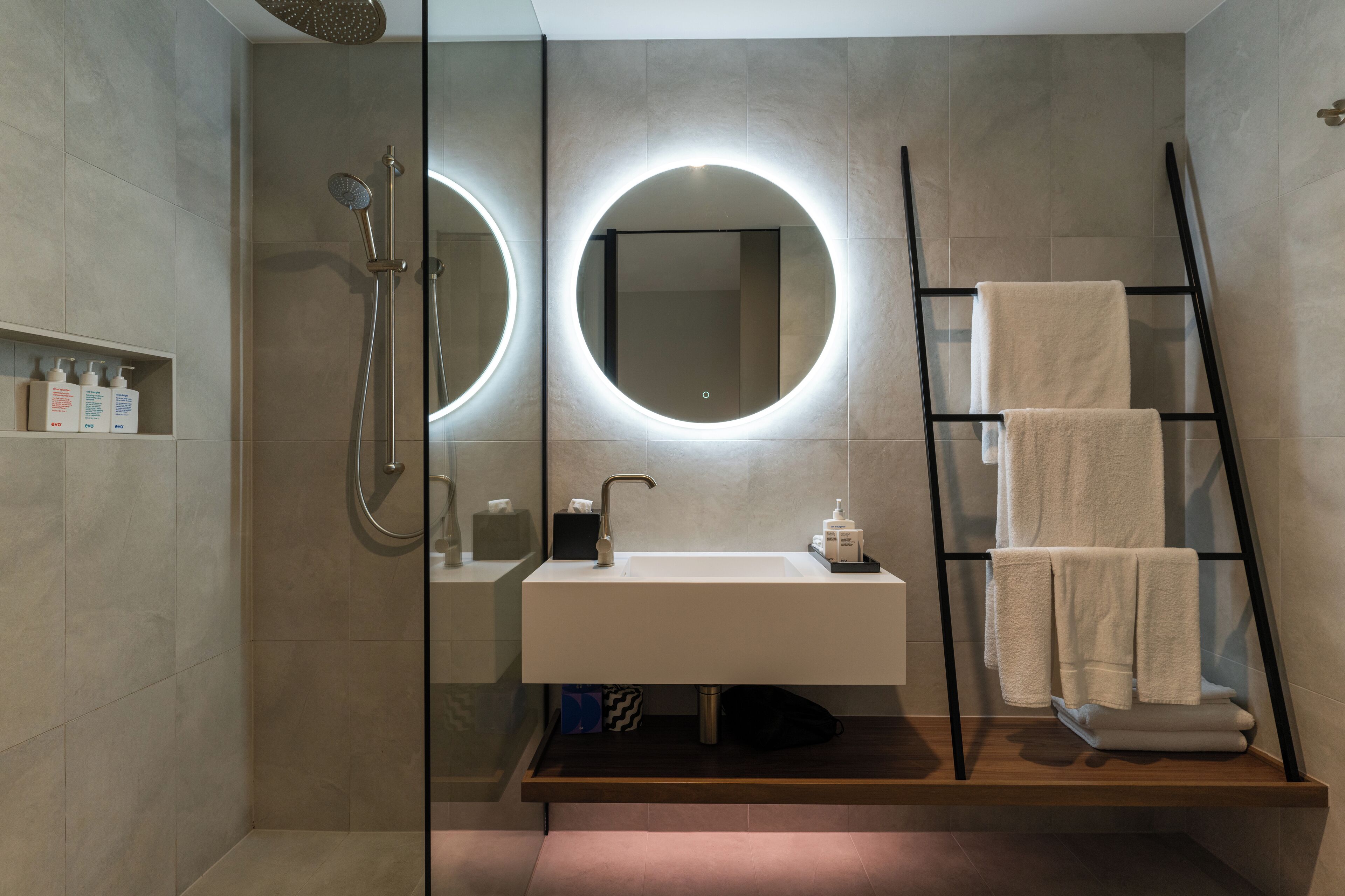Ein modernes Badezimmer mit einer einachigen Duschkabine, einem runden Spiegel, einem Waschbecken mit Handtuchhalter und einem Holzregal.