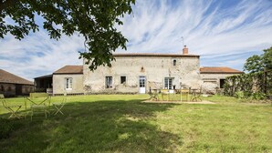 A rustic stone building with a tiled roof, a grassy yard, and a small table with chairs.