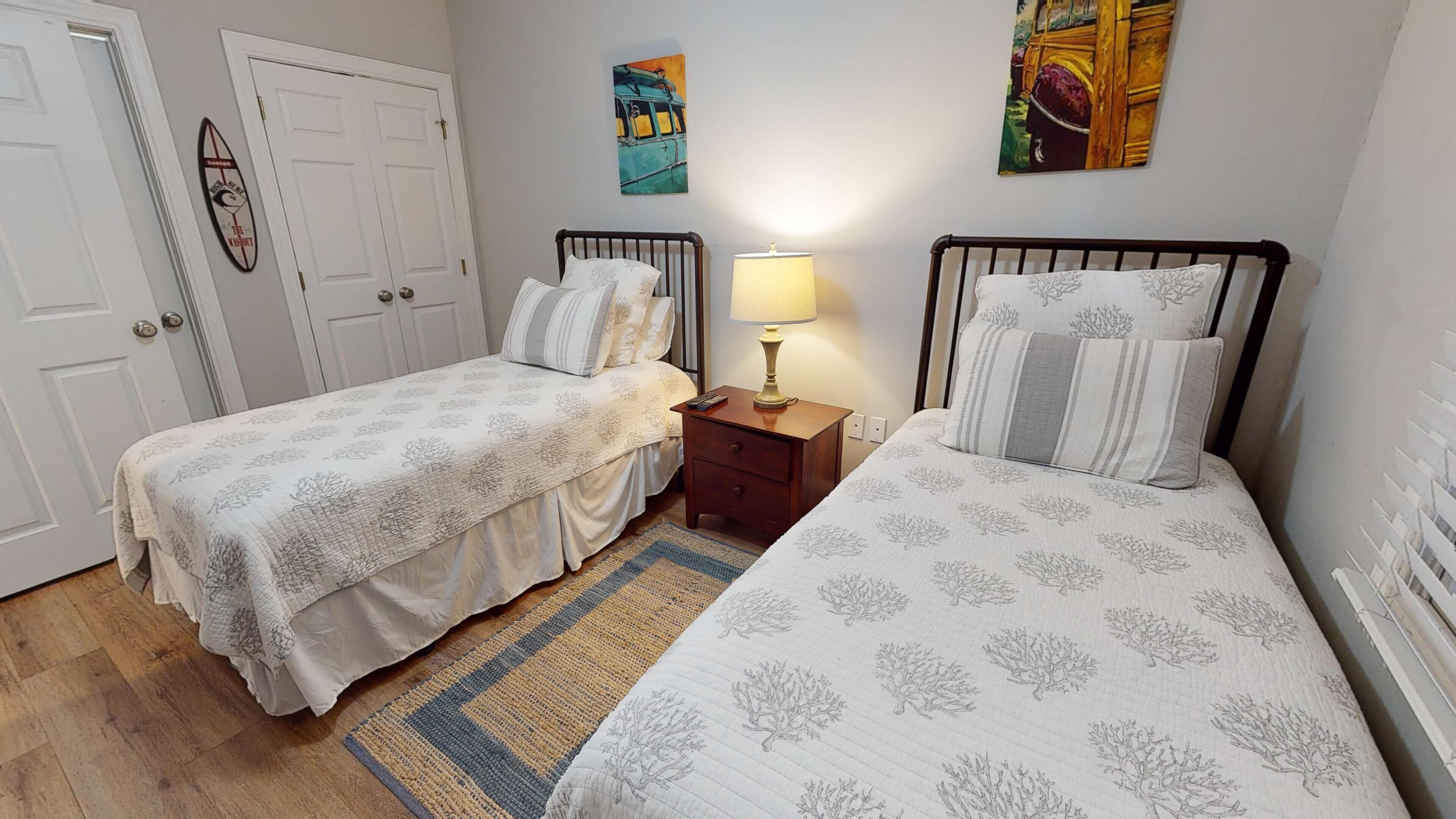A bedroom with two single beds, a nightstand with a lamp, a door, and two paintings on the wall.