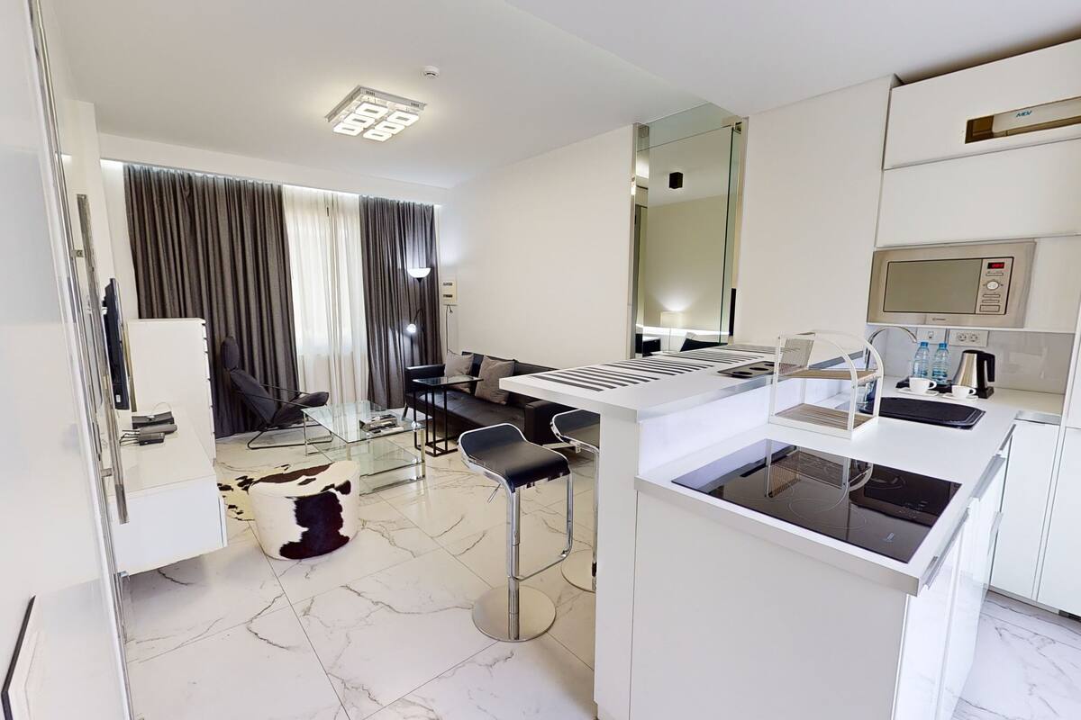 Una habitación de hotel moderna con kitchenette, una zona de estar con sofá y sillones, y un espejo grande.