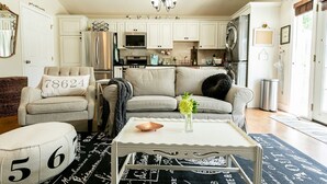 A living room with a sofa, armchair, and ottoman, a coffee table with a vase of flowers, and a kitchen in the background.