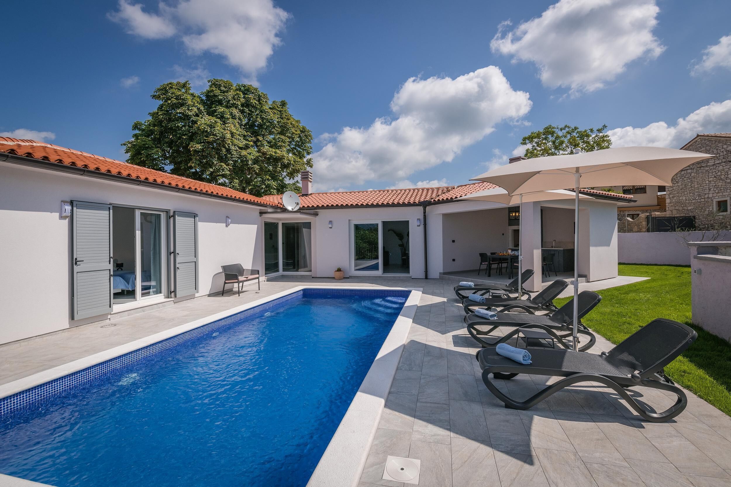 A swimming pool with lounge chairs and a covered seating area, surrounded by a modern building with a tiled roof.