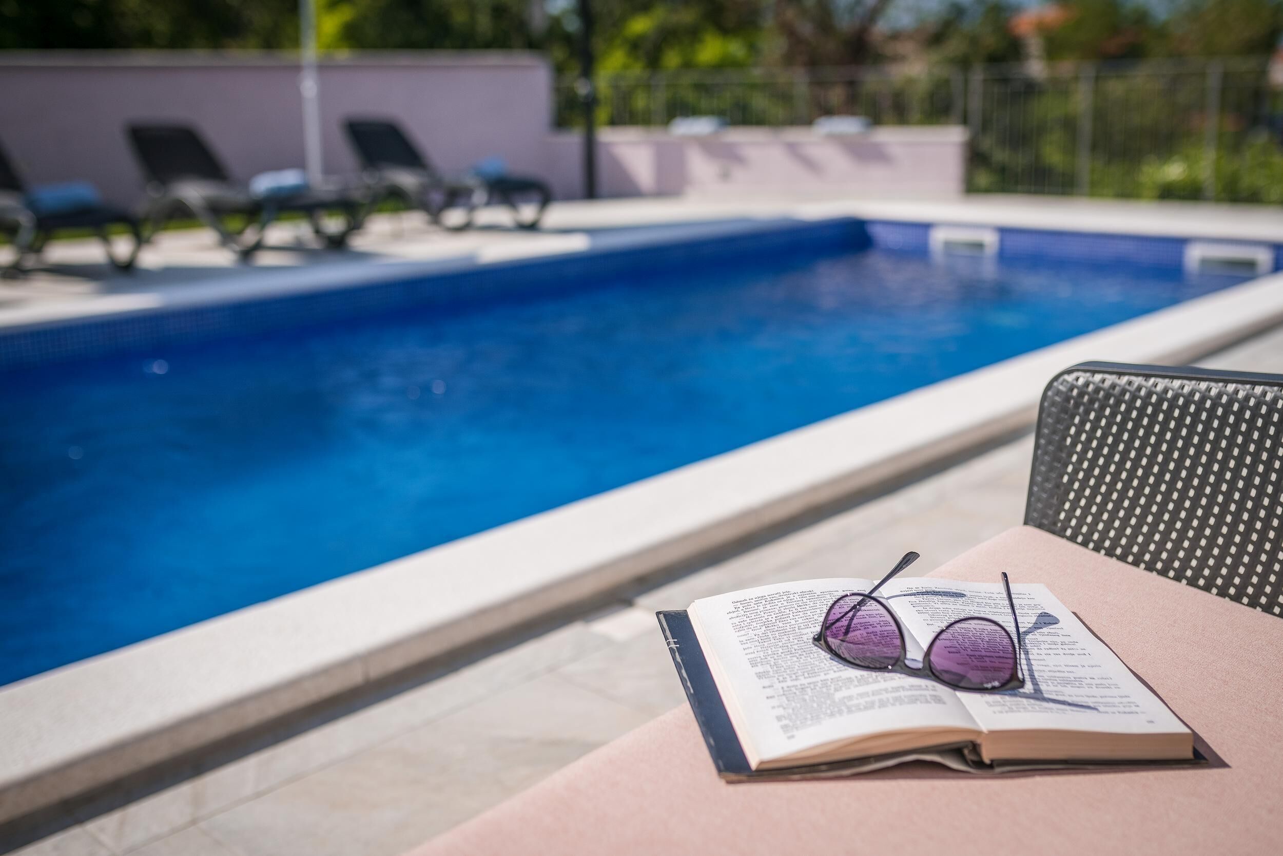 Une scène au bord de la piscine, avec un livre et des lunettes posés sur une table, et des chaises longues à l’arrière-plan.
