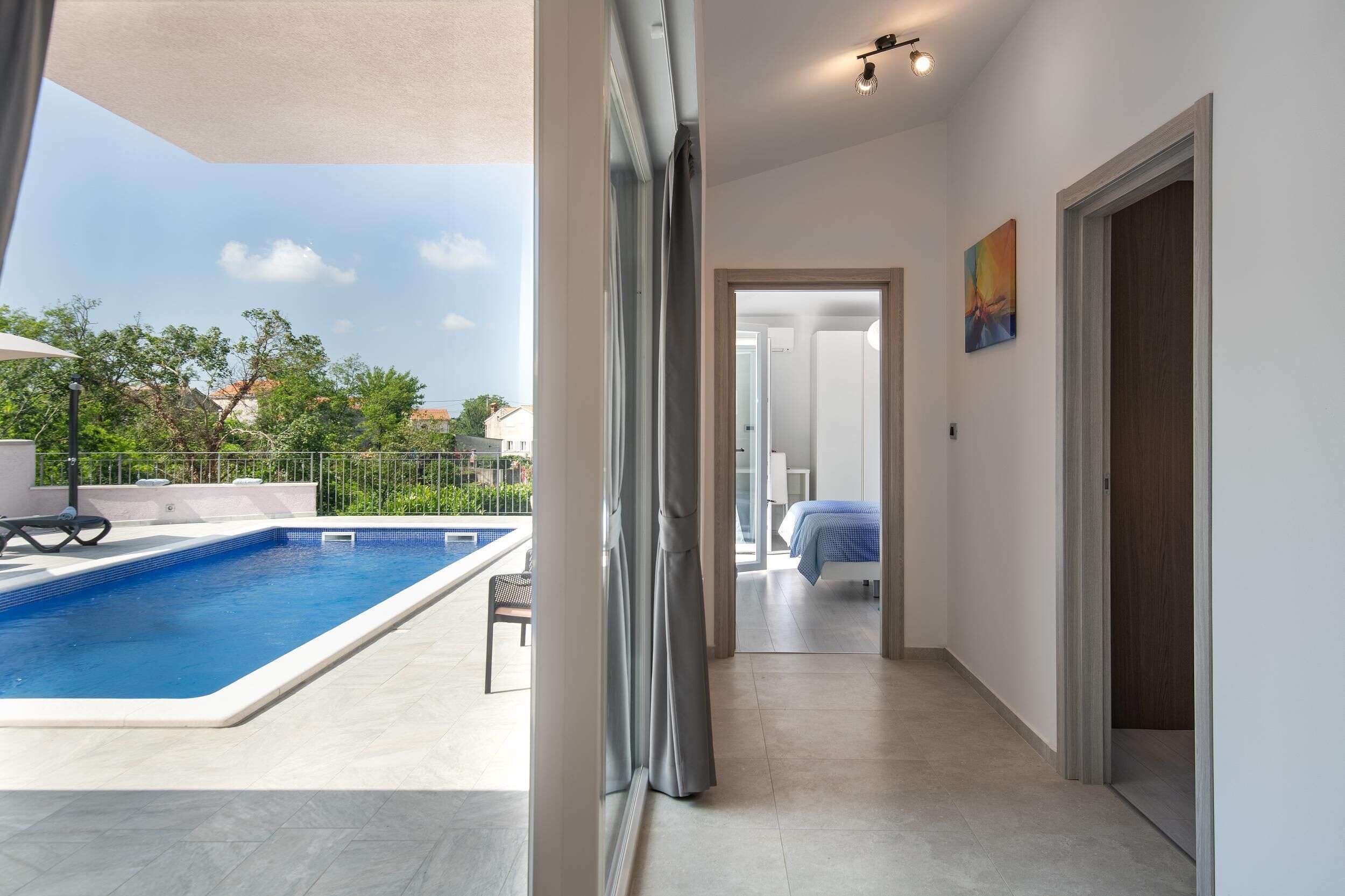 A modern hotel room with a view of a swimming pool, a balcony with a chair, and a door leading to another room.