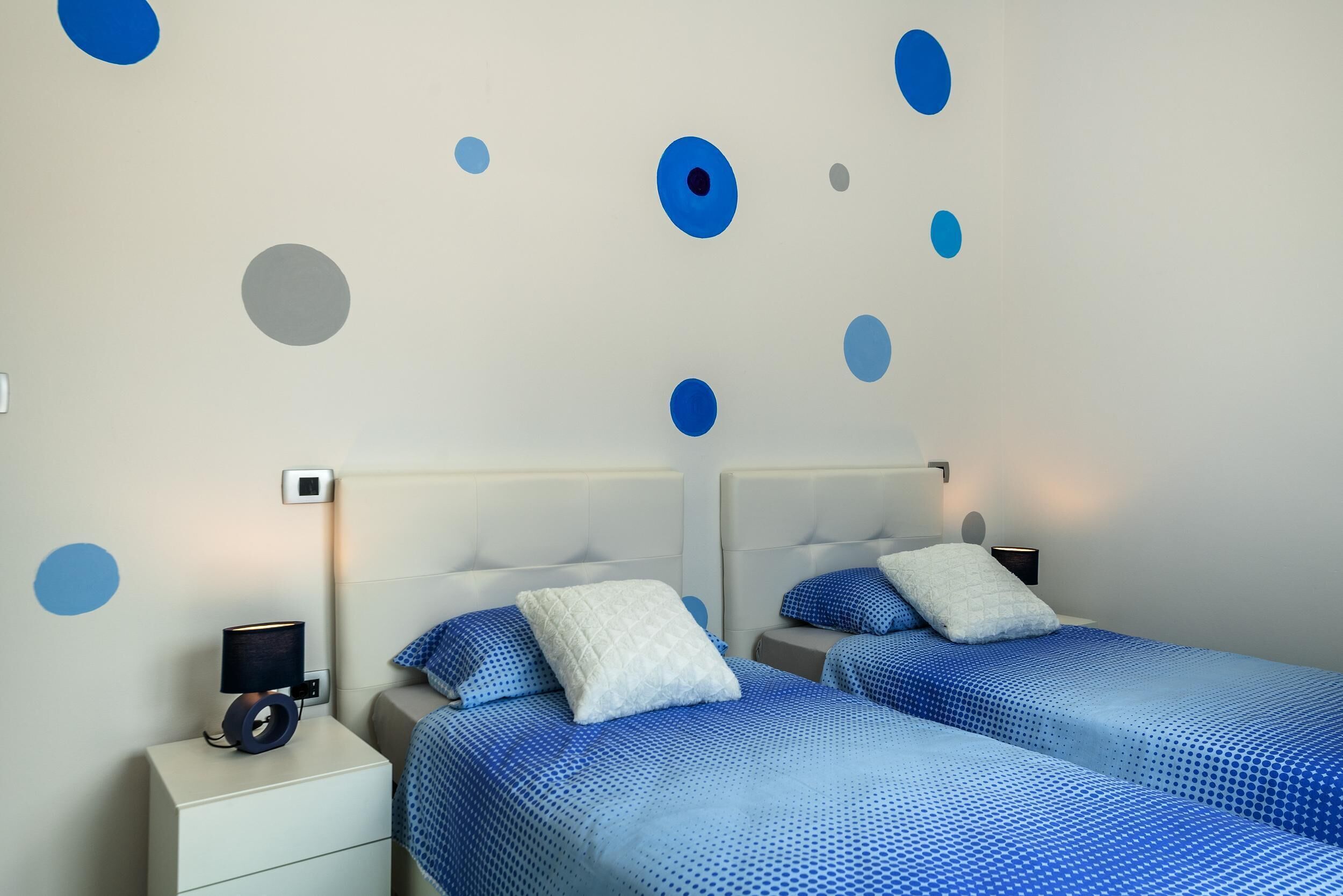 A bedroom with two beds, each with blue bedding and white pillows, a nightstand with a lamp, and a wall with blue and gray circular decorations.
