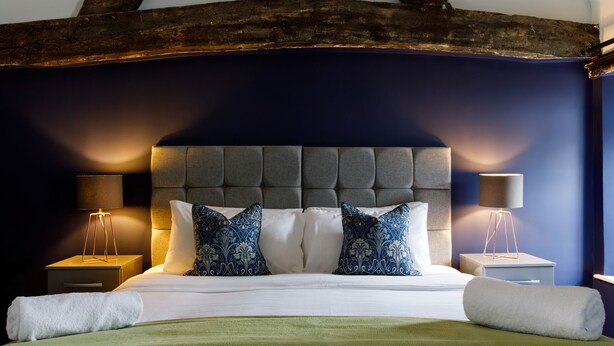 A neatly made bed with white linens and blue patterned pillows, flanked by two bedside tables with lamps, in a room with dark blue walls and exposed wooden beams. - Canon Gate , Chichester Cathedral  - sleeps 4 guests  in 2 bedrooms (Chichester)