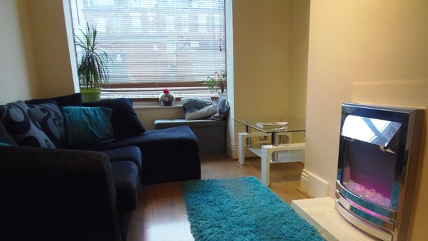 A living room with a dark-colored sofa, a teal rug, a glass-top coffee table, and a flat-screen TV. - Light and Airy 1 bed Flat With Large Basement Kitchen and Private Outside Area (leeds)