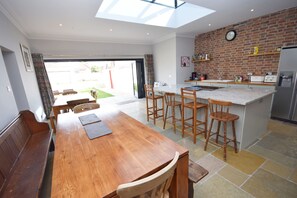 A spacious kitchen with a large island, wooden dining table, and chairs. There is a brick wall and a clock on the wall.