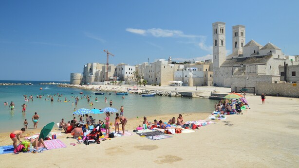 Uma praia com pessoas tomando banho de sol e nadando, um edifício histórico com torres, e um céu azul e límpido.