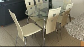 A dining area with a glass table and chairs, a ceiling fan, an air conditioning unit, and a dark-colored sofa with a red cushion. - KINGS SUNSET, PAPHOS (Paphos)