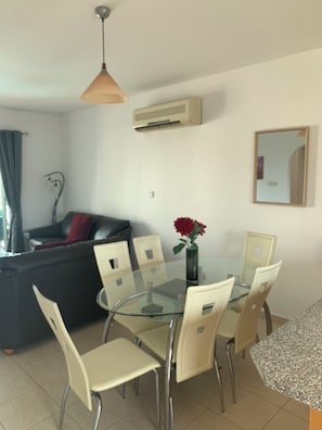 A dining area with a glass table and chairs, a ceiling fan, an air conditioning unit, and a dark-colored sofa with a red cushion. - KINGS SUNSET, PAPHOS (Paphos)