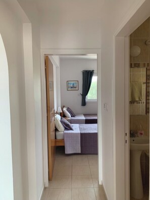 A bedroom with two beds, a window with curtains, and a bathroom with a sink and mirror. - KINGS SUNSET, PAPHOS (Paphos)