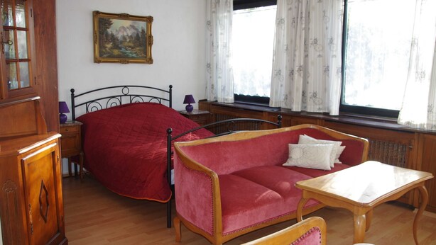 Ein Zimmer mit einem Holzkabinett, einem roten Sofa, einem kleinen Tisch und einem Fenster mit Vorhängen.