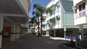 A multi-story hotel with a central courtyard, featuring a paved walkway, palm trees, and a bench. - 222, apartment in a gated community with a shared swimming pool. (Bombas)
