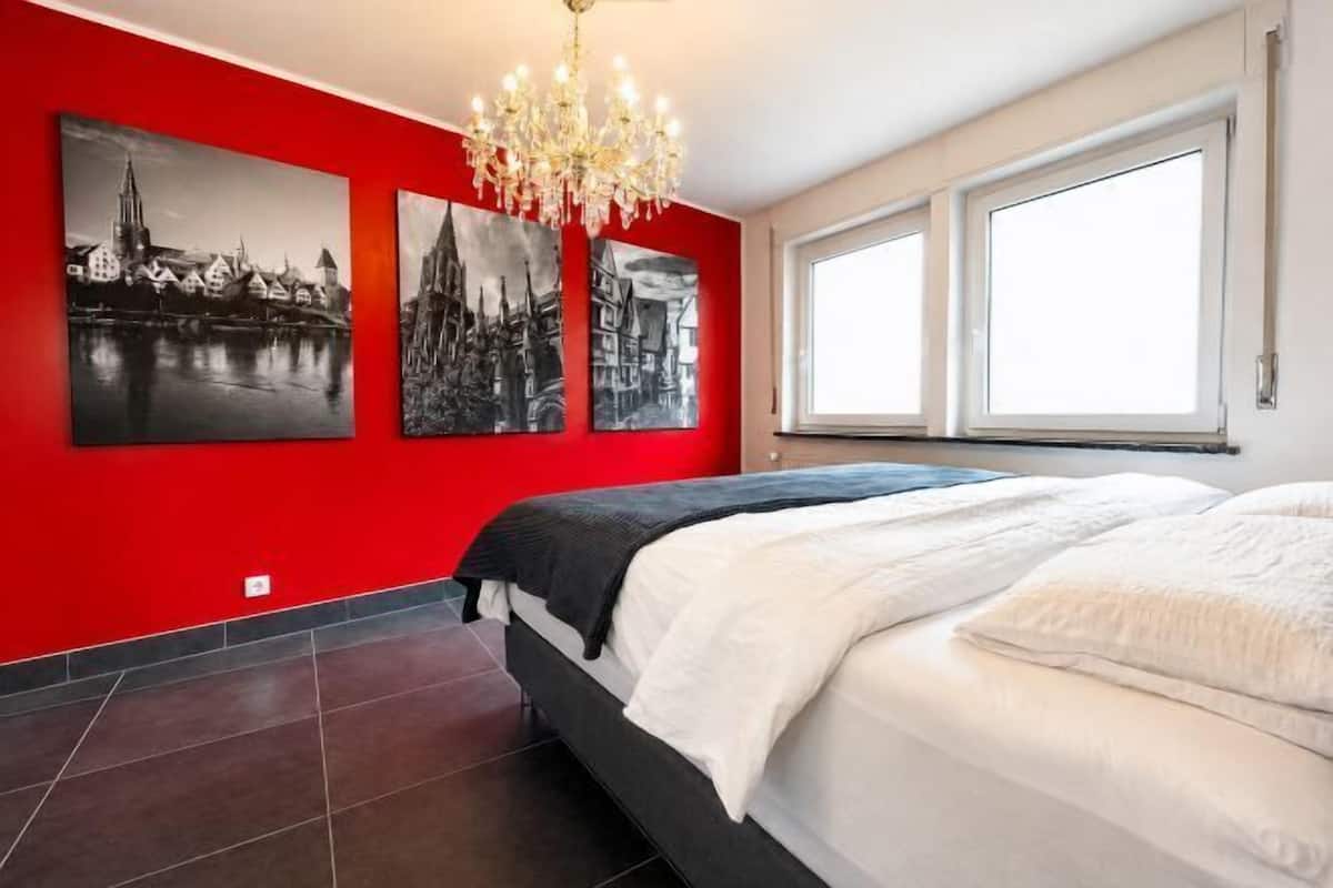 Un dormitorio con una cama grande, una araña de cristal y tres pinturas en blanco y negro de paisajes urbanos en la pared roja.