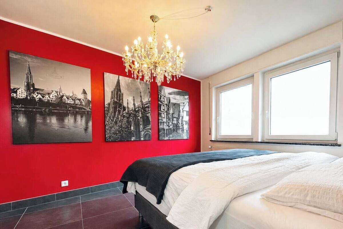 Un dormitorio con una cama, una araña y tres pinturas en blanco y negro de un paisaje urbano sobre una pared roja.
