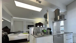 Une chambre d’hôtel moderne équipée d’une kitchenette, d’un lit avec un oreiller vert, d’une salle de bain avec des toilettes et d’un téléviseur fixé au mur.