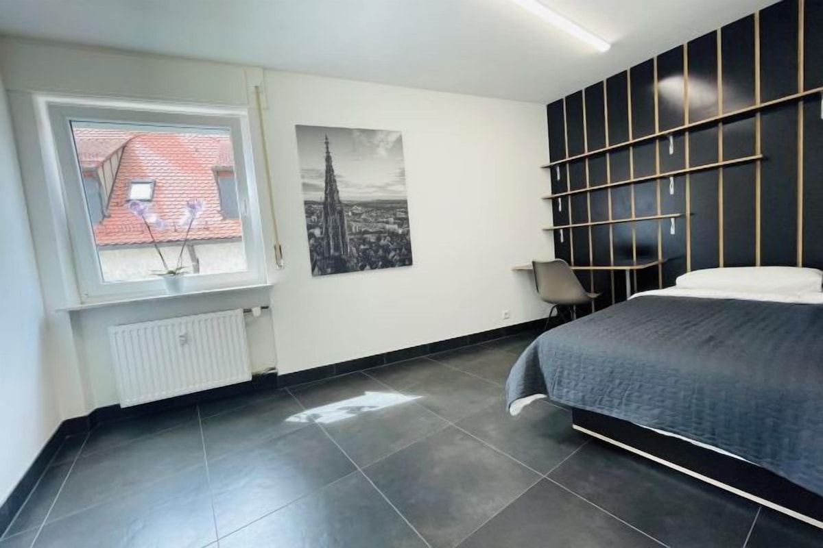 A modern bedroom with a large bed, a window with a view of a building, a radiator, and a wall-mounted shelf unit.
