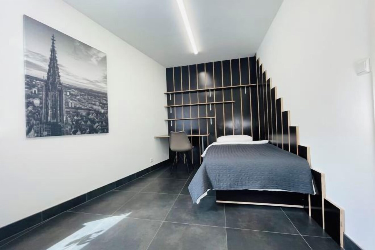 A modern bedroom with a bed, a chair, a desk, and a cityscape painting on the wall.