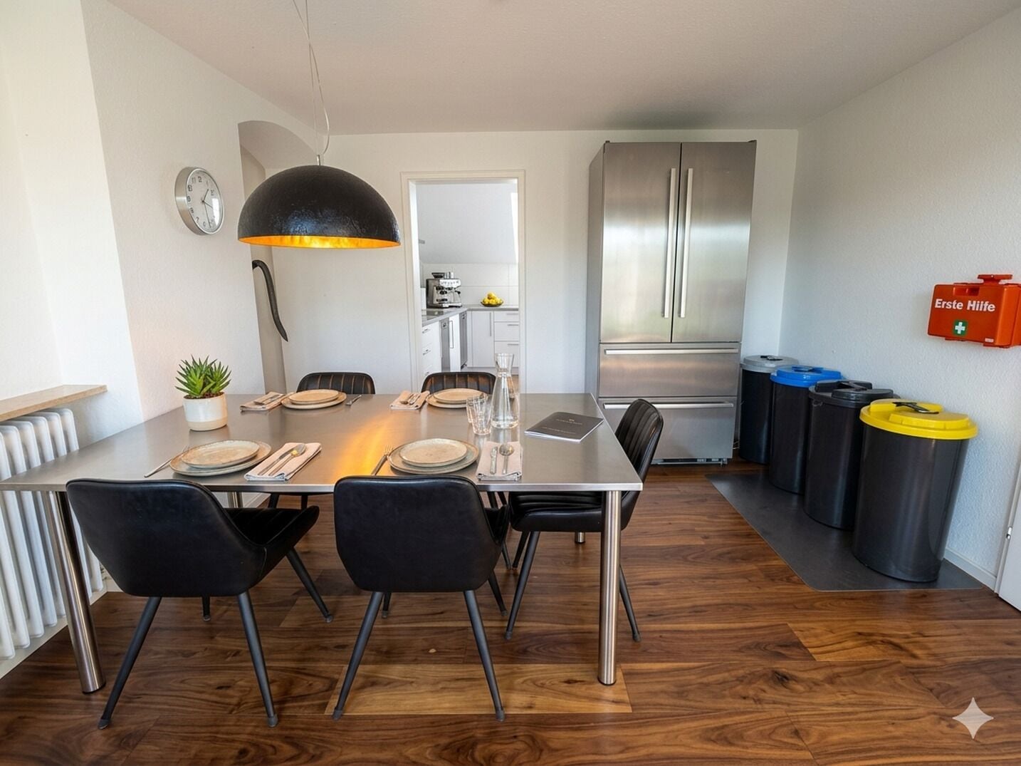 Uma cozinha moderna com eletrodomésticos de aço inoxidável, uma área de jantar com mesa e cadeiras de madeira e um cesto de lixo com tampas coloridas.