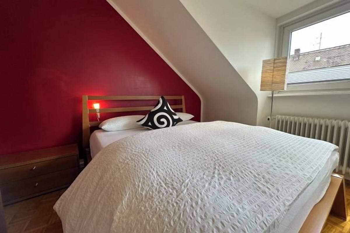 A bedroom with a sloped ceiling, a bed with a white bedspread, a red wall, and a window with blinds.
