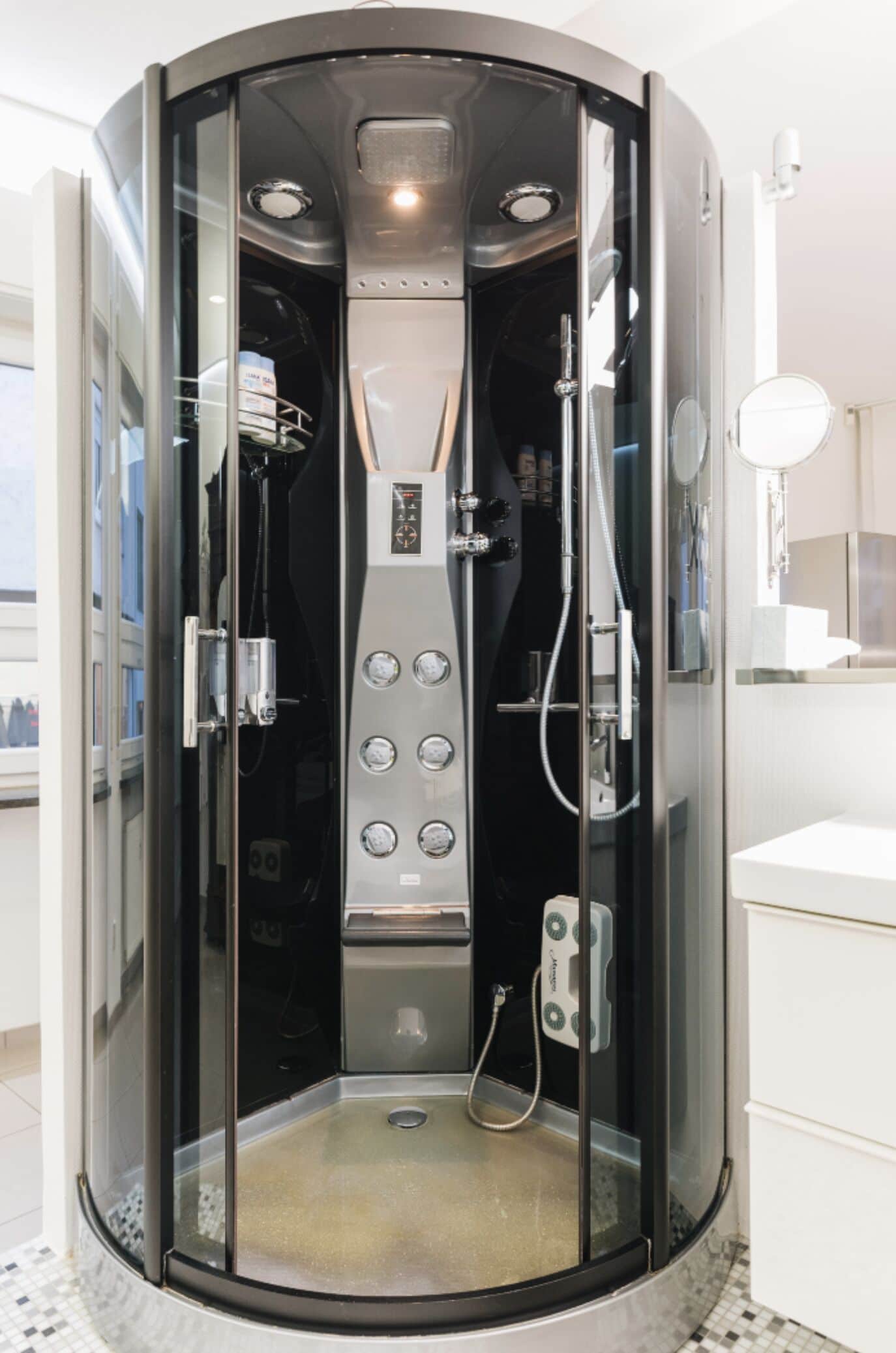 A modern, enclosed shower stall with a control panel, a handheld showerhead, and a handheld vacuum.
