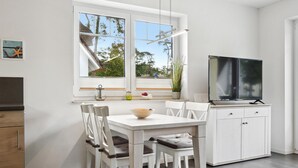 Moderni keittiö, jossa on valkoiset kaapit, ruokapöytä tuoleineen, televisio ja ikkuna, josta avautuu näkymä puita kohti.