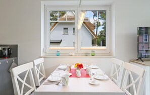 Un comedor con una mesa dispuesta para cuatro personas, sillas blancas, una ventana con vistas a una casa y un televisor sobre un mueble.
