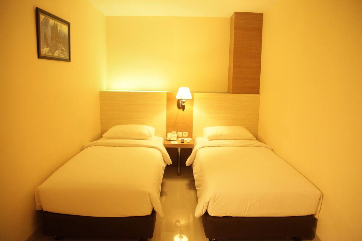 Une chambre d’hôtel avec deux lits simples, une table de chevet avec une lampe, un téléphone et une petite table avec une horloge.
