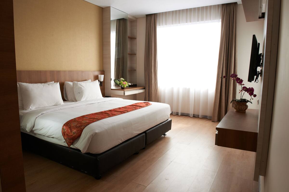 Une chambre d’hôtel avec un grand lit, une table de chevet ornée d’un vase de fleurs, un téléviseur à écran plat fixé au mur et une grande fenêtre avec des rideaux.