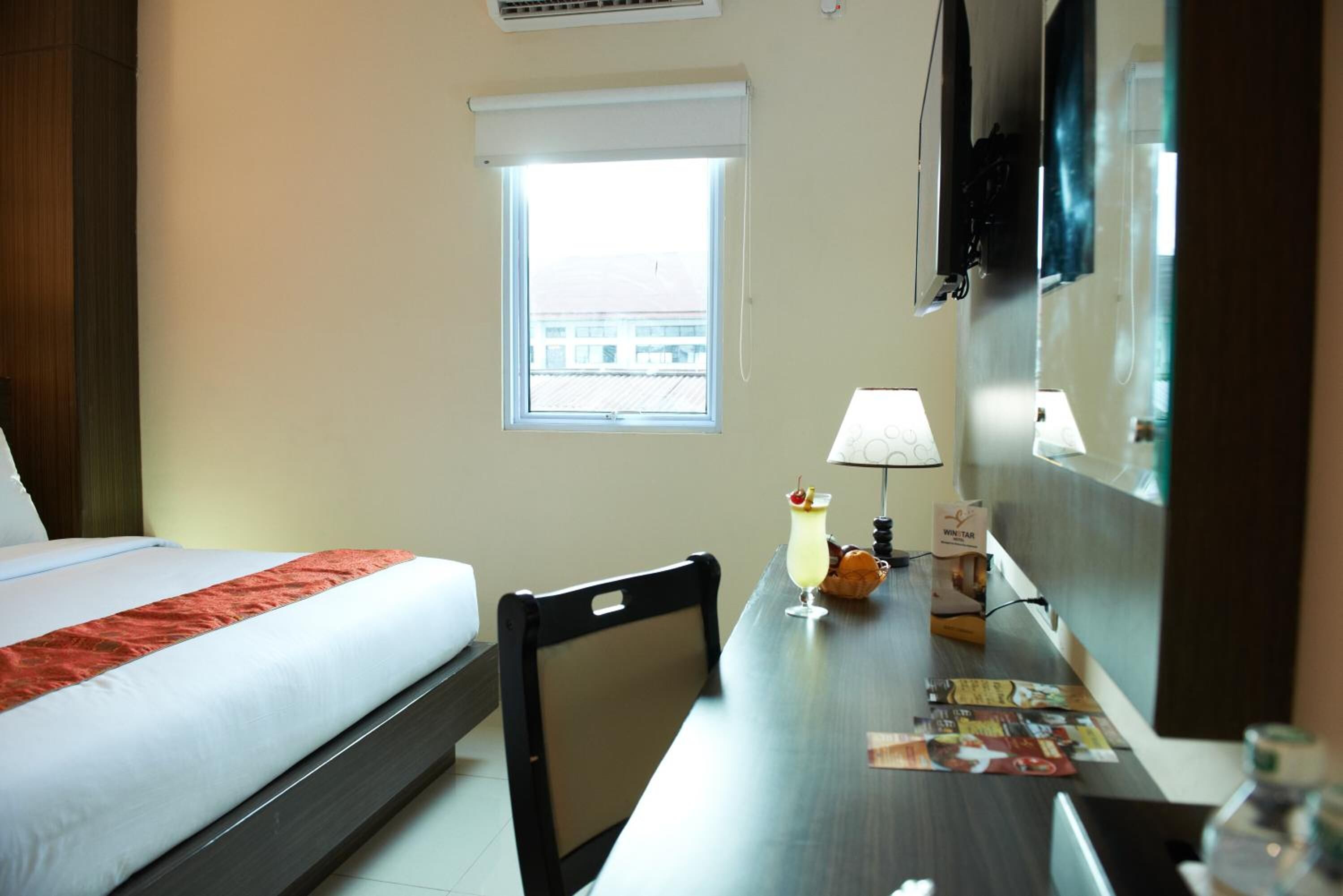 A hotel room with a bed, a desk with a lamp, a window, and a view of the outside.