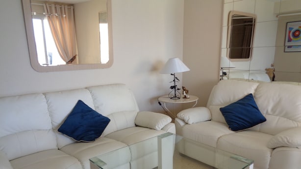 A living room with two white sofas, each with a blue pillow, a glass coffee table, a small white side table with a lamp, and a mirror on the wall. - A63 PEÑON DE IFACH FASE1 9ºD (Calpe)