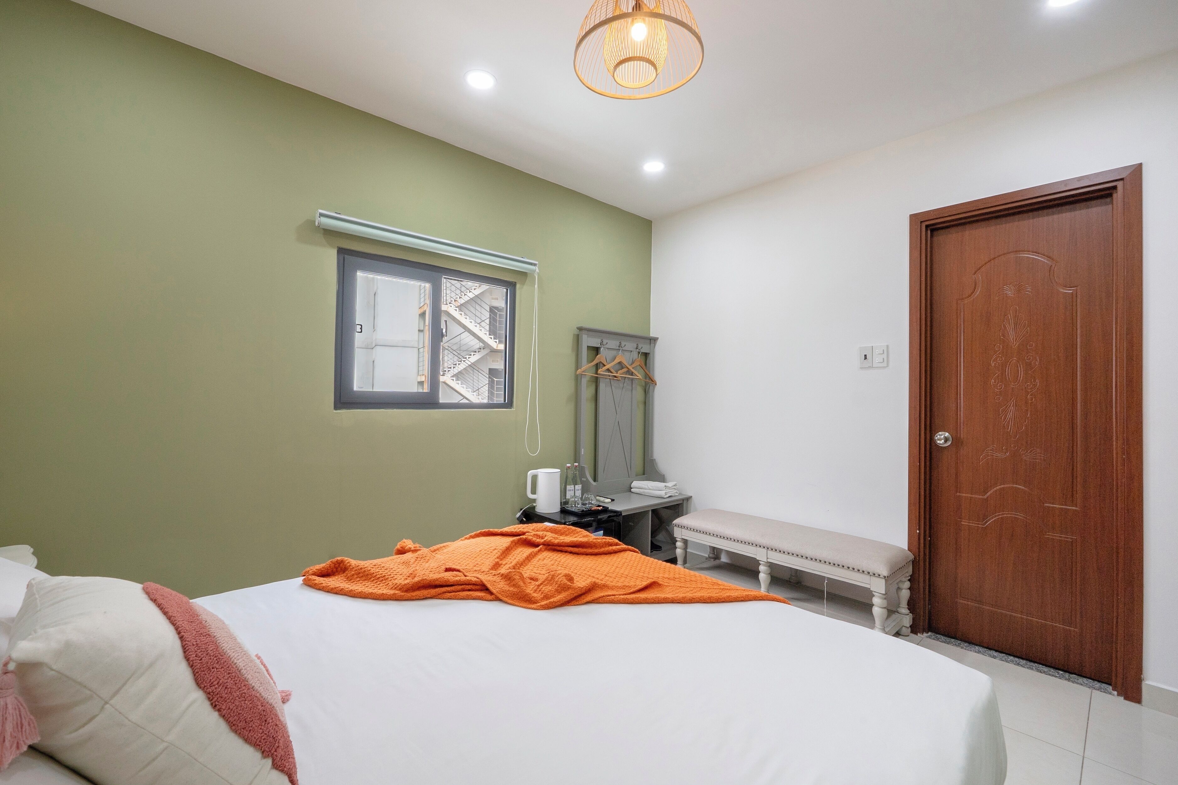 A bedroom with a bed, a bench, a wooden door, a window with a view of a building, and a hanging coat rack.