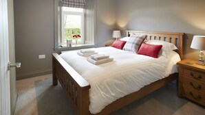Un lit bien fait, avec du linge de lit blanc et un coussin rouge, une table de chevet en bois avec une lampe, et une fenêtre donnant sur un paysage verdoyant.