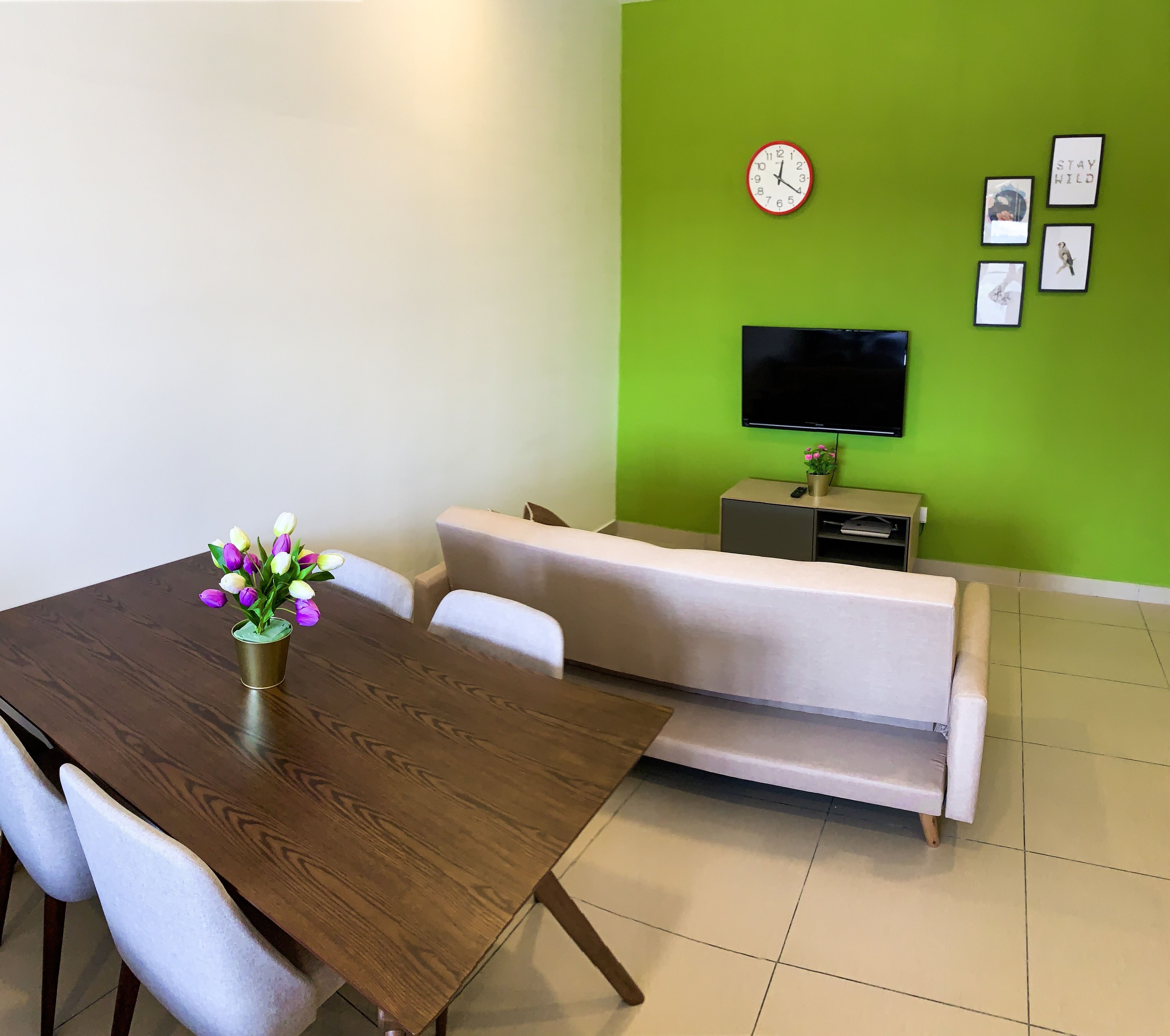 A modern living room with a wooden dining table, a sofa, a TV, and a green accent wall.