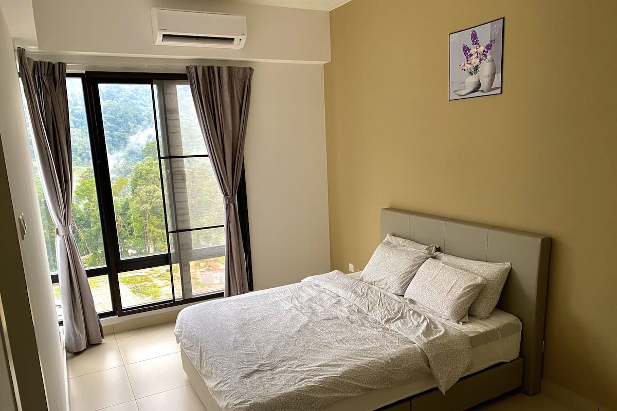 A bedroom with a bed, curtains, an air conditioning unit, and a window with a view of greenery.