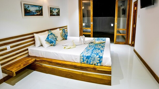 A bedroom with a wooden bed frame, white bedding, and a blue floral throw. There are two paintings on the wall and a sliding glass door leading to an outdoor area. - Ricco Tara (Boossa)