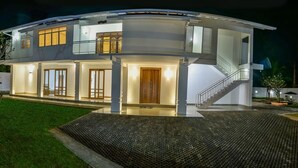 A modern two-story house with a covered patio, a staircase, and a well-lit entrance. - Ricco Tara (Boossa)