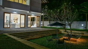 A modern two-story house with a well-lit interior, a balcony, and a landscaped backyard with a tree and outdoor furniture. - Ricco Tara (Boossa)