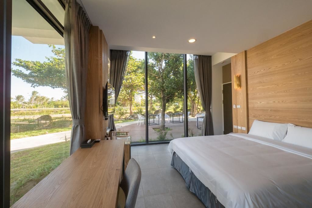 A modern hotel room with a large bed, a wooden desk, and a view of a garden and street.