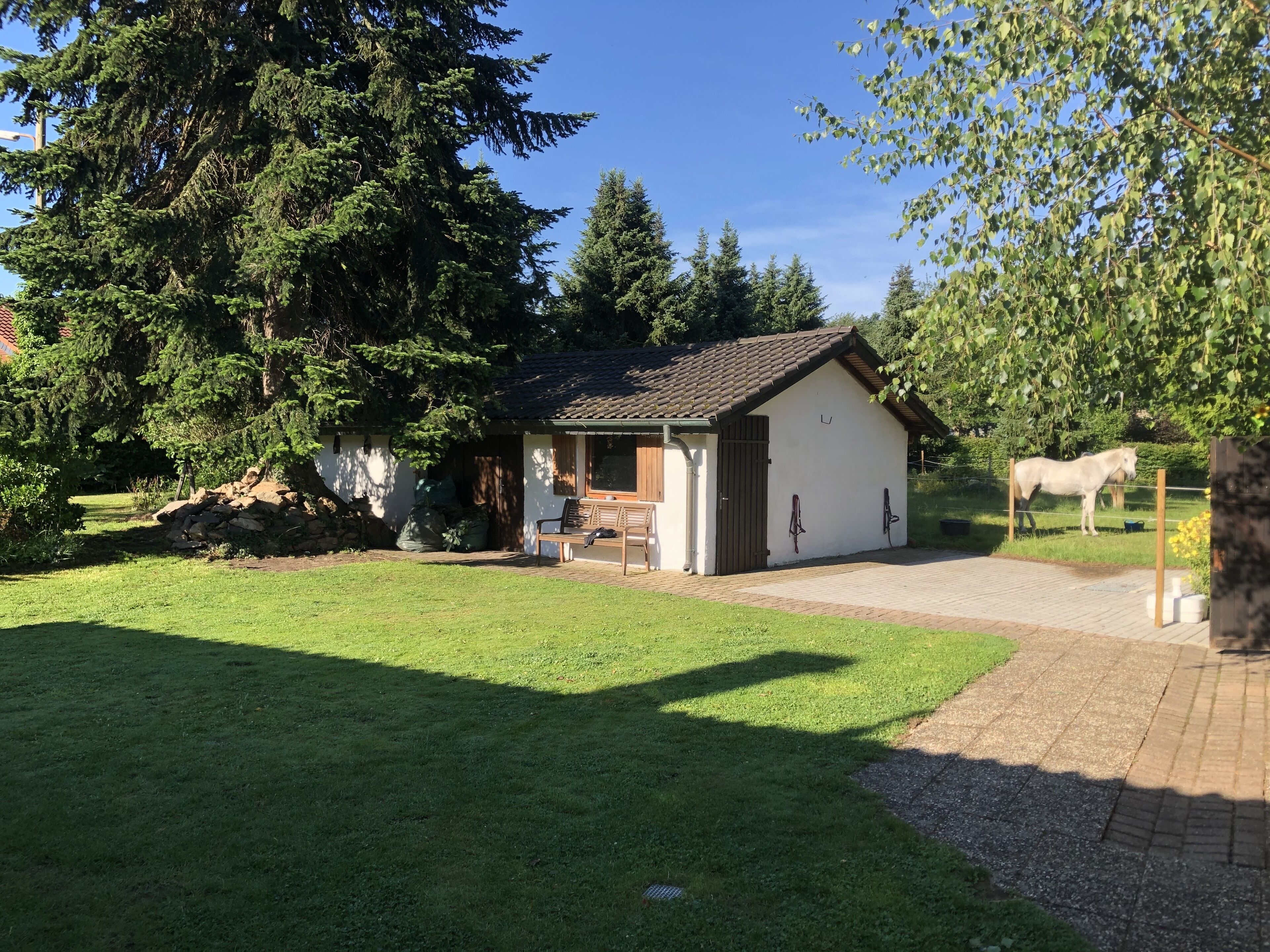 A small, single-story building with a tiled roof, a covered porch, and a fenced area with a horse.