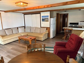 Ein Wohnzimmer mit einer Couch, einem Holztisch und einem roten Sessel. Es gibt eine Küchenzeile mit einem Kühlschrank und einer Mikrowelle.