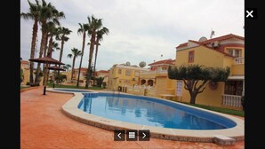 A swimming pool with a tiled deck, surrounded by palm trees and buildings with Mediterranean architecture. - Air-conditioned 2 bedroom villa in Orihuela Costa with pool and sea 1 km (Orihuela Costa)