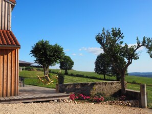 A wooden building with a tiled roof, a wooden deck, and a stone wall with a flower bed. - The ash trees of Augustus (Clavières)