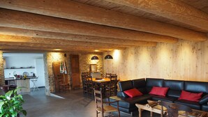 A cozy interior with a wooden ceiling, a black leather sofa with red cushions, a dining area with a wooden table and chairs, and a kitchen with stone walls and wooden beams. - The ash trees of Augustus (Clavières)
