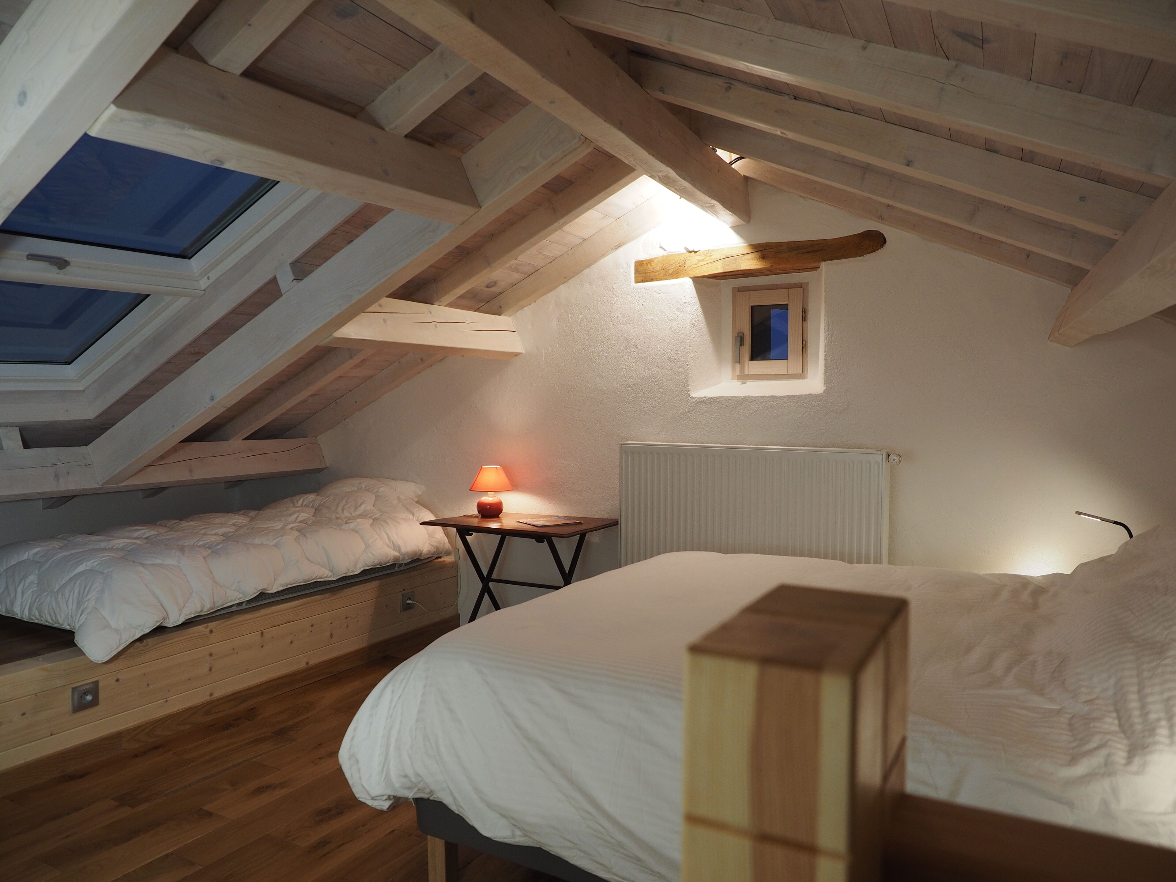 Ein Schlafzimmer mit schrägen Wänden, Holzbalken, einem Oberlicht, einem kleinen Tisch mit Lampe und zwei Betten.