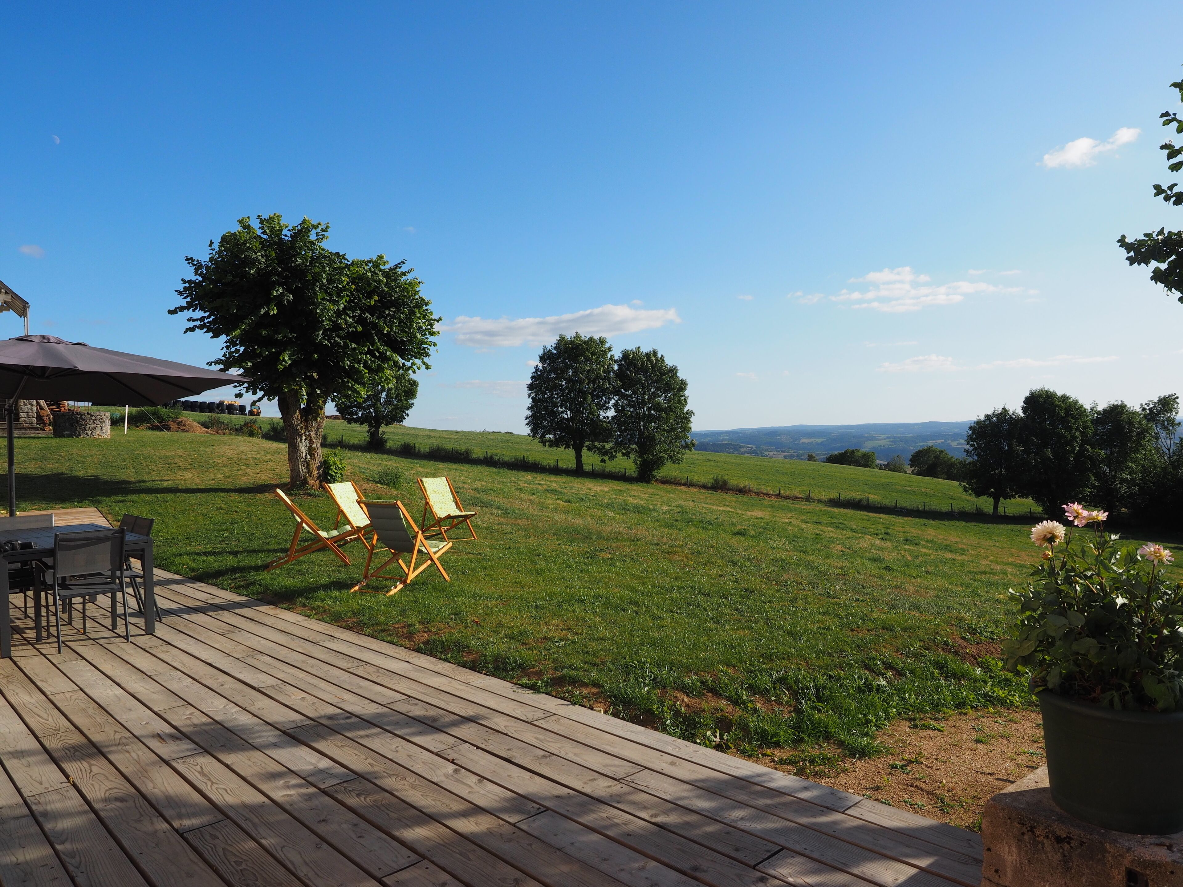 A wooden deck with outdoor furniture, a table, and chairs, overlooking a grassy landscape with trees and a clear sky.