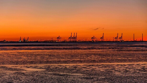Ein Sonnenuntergang über einer Küstenlandschaft mit Industriegebäuden, wobei Kräne und Bauwerke am Himmel in Silhouette zu sehen sind.