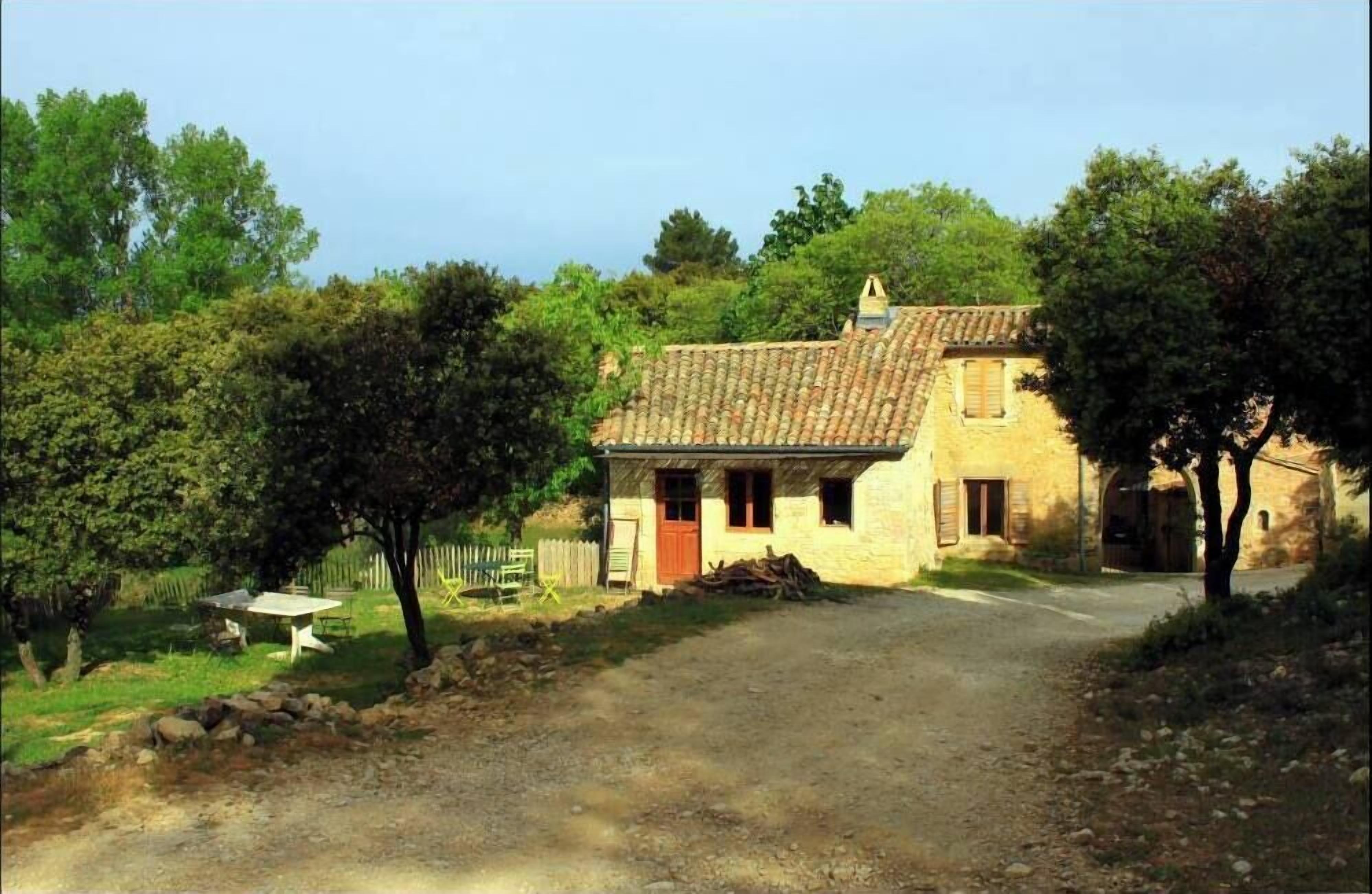 Una casa de piedra rústica con techo de tejas, rodeada de árboles y un camino de grava.