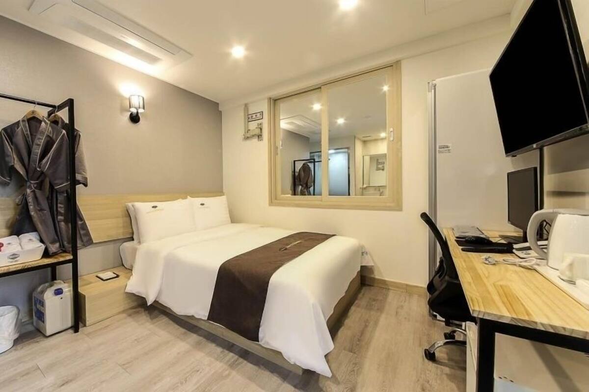 A hotel room with a bed, a desk with a computer and a coffee maker, a mirror, and a coat rack with clothes.